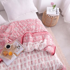 May include: A pink and white paisley patterned quilt with a matching pillow on a bed. A small wooden side table with a book and flowers sits next to the bed. A tray with coffee, a pastry, and an orange slice rests on the quilt.