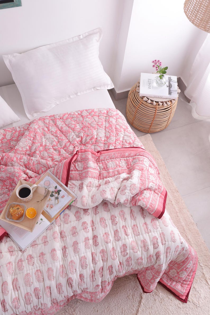 May include: A pink and white paisley patterned quilt with a matching pillow on a bed. A small wooden side table with a book and flowers sits next to the bed. A tray with coffee, a pastry, and an orange slice rests on the quilt.