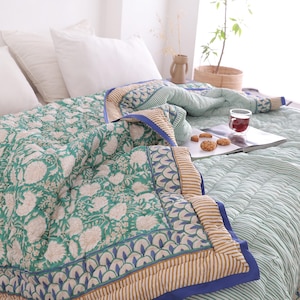 Op de afbeelding: Een bed is bedekt met een groen bloemenpatroon quilt en witte kussens. De quilt heeft een blauwe rand met een beige en wit gestreept patroon. Een glas thee en koekjes liggen op een tijdschrift.