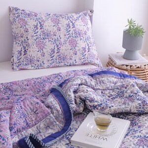 May include: A floral patterned quilt and pillow set in shades of purple and blue. The bedding features a repeating flower design on a white background. A cup of tea sits on a book with the word "WHITE" on it.