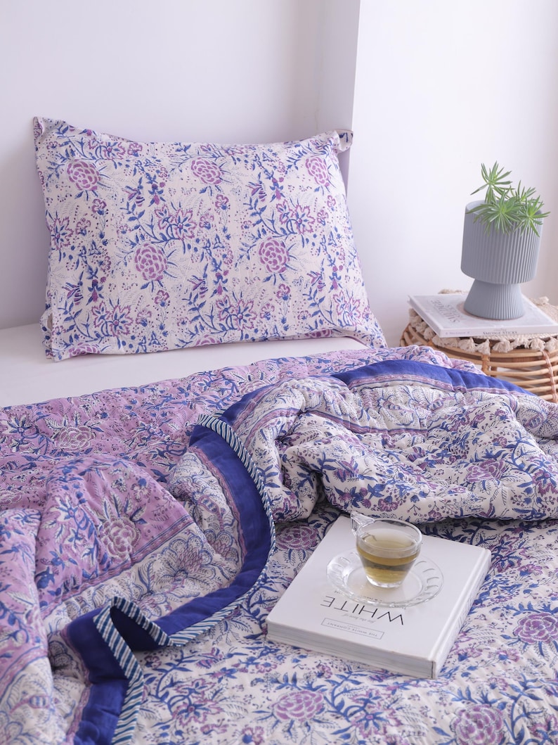 May include: A floral patterned quilt and pillow set in shades of purple and blue. The bedding features a repeating flower design on a white background. A cup of tea sits on a book with the word "WHITE" on it.