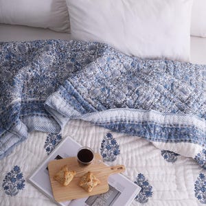 May include: A bed is made with white pillows and a blue and white floral patterned quilt. A wooden cutting board with pastries and a glass of dark liquid sits on a magazine.