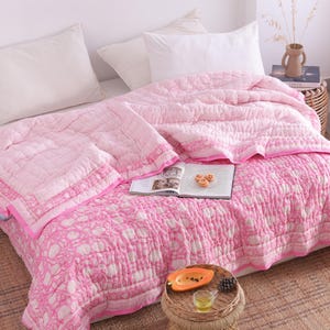 May include: A pink and white patterned quilt and matching pillow set on a bed. The quilt features a floral design and a solid pink border. The bed is made up with white pillows and a woven rug is on the floor.