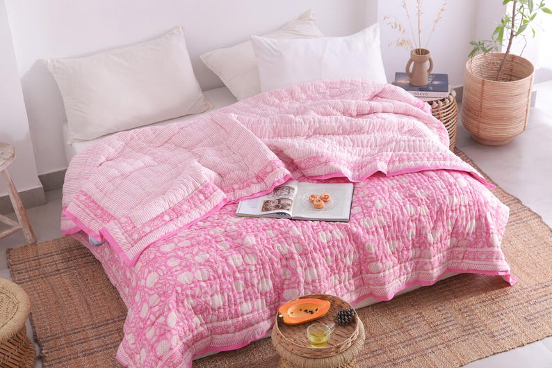May include: A pink and white patterned quilt and matching pillow set on a bed. The quilt features a floral design and a solid pink border. The bed is made up with white pillows and a woven rug is on the floor.