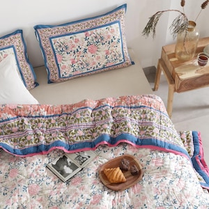 May include: A floral patterned quilt and matching pillow set in pink, blue, and white. The quilt features a repeating floral design with a blue border and pink trim. Two pillows with a similar floral pattern are visible. A wooden side table with a candle and vase is in the background.