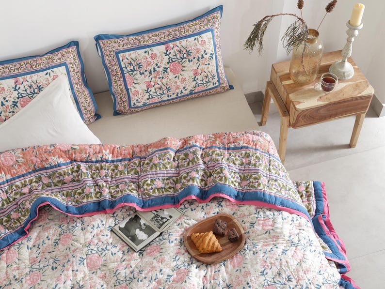 May include: A floral patterned quilt and matching pillow set in pink, blue, and white. The quilt features a repeating floral design with a blue border and pink trim. Two pillows with a similar floral pattern are visible. A wooden side table with a candle and vase is in the background.