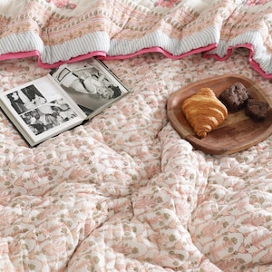 Puede incluir: Un edredón acolchado con un estampado floral en tonos rosas y blancos. Una bandeja de madera con un cruasán y dos magdalenas de chocolate. Un libro abierto con fotos en blanco y negro descansa sobre el edredón.