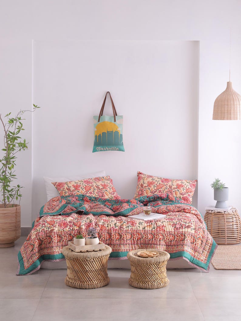 May include: A bedroom scene featuring a floral patterned quilt and matching pillows in shades of red, pink, and teal. Two small woven stools sit at the foot of the bed. A tote bag with a Nashville skyline hangs on the wall.