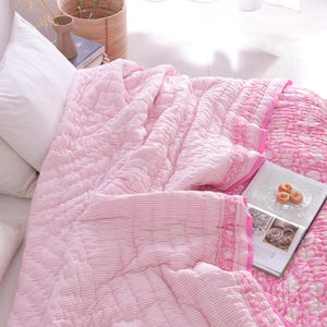 May include: A pink and white patterned quilt on a bed with white pillows. The quilt has a striped design on one side and a floral pattern on the other. A book and pastries are on the quilt, creating a cosy bedroom scene.