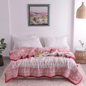 May include: A bed is covered with a pink and white patterned quilt. The quilt has a floral design and a red border. Two white pillows rest against the wall. A framed landscape painting hangs above the bed.