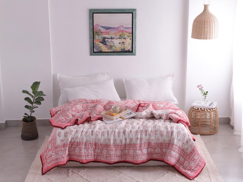May include: A bed is covered with a pink and white patterned quilt. The quilt has a floral design and a red border. Two white pillows rest against the wall. A framed landscape painting hangs above the bed.