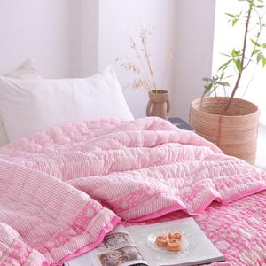 May include: A pink and white patterned quilt and pillow set on a bed. The quilt features a striped design with floral detailing. A book and pastries are on the quilt. A woven basket with a plant is in the background.