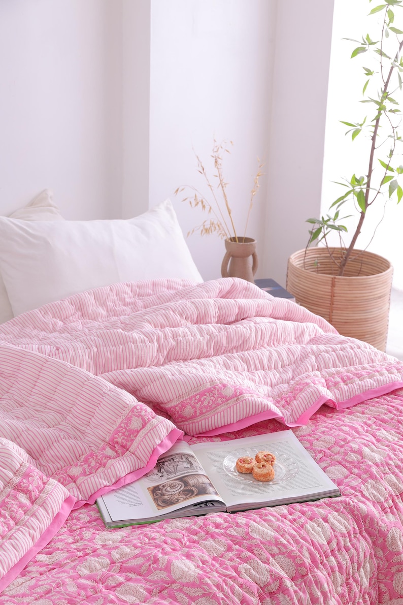 May include: A pink and white patterned quilt and pillow set on a bed. The quilt features a striped design with floral detailing. A book and pastries are on the quilt. A woven basket with a plant is in the background.
