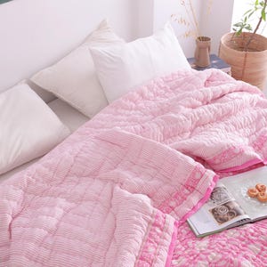 May include: A bed with white pillows and a pink quilted blanket. The blanket has a striped pattern on one side and a floral pattern on the other. A book and small pastries are on the bed.