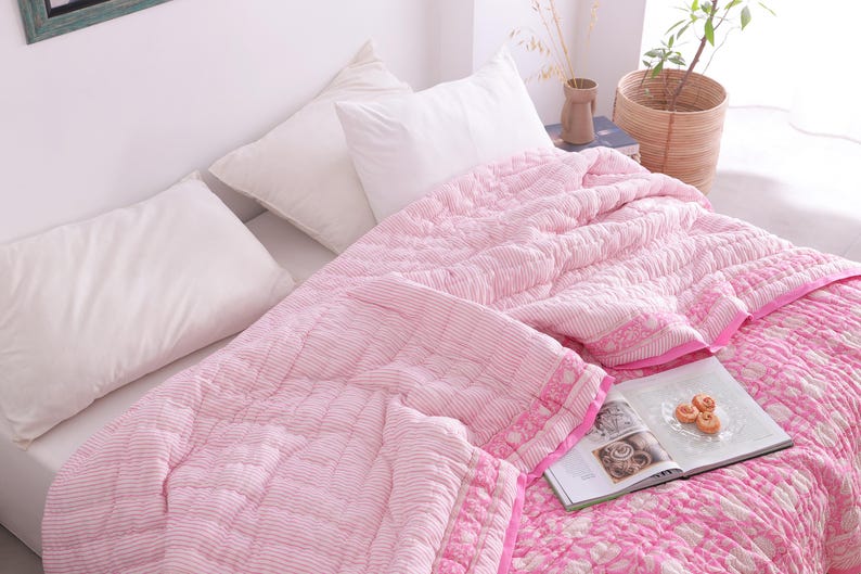 May include: A bed with white pillows and a pink quilted blanket. The blanket has a striped pattern on one side and a floral pattern on the other. A book and small pastries are on the bed.