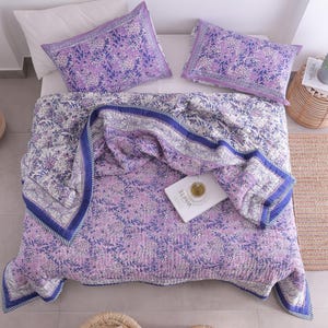 May include: A bed is made with a floral patterned quilt and two matching pillowcases in shades of purple and white. The quilt has a blue border. A book with the word "WHITE" is on the quilt. A small woven side table and a woven rug are also in the image.