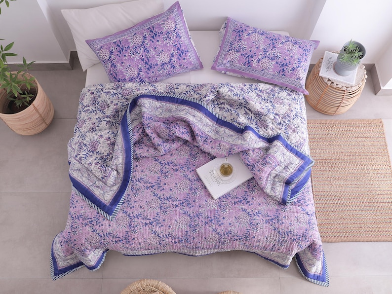 May include: A bed is made with a floral patterned quilt and two matching pillowcases in shades of purple and white. The quilt has a blue border. A book with the word "WHITE" is on the quilt. A small woven side table and a woven rug are also in the image.
