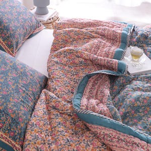 May include: A floral patterned quilt and two matching cushions in teal, pink, and yellow. The quilt has a teal border. A book and a cup of tea sit on the quilt. The cushions have a teal border.