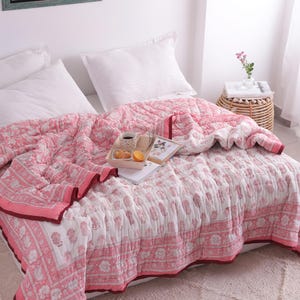 May include: A pink and white floral patterned quilt covers a bed with white pillows. The quilt has a decorative border and is paired with a tray holding a cup, pastries, and an orange slice. A small side table with a vase of flowers is visible.
