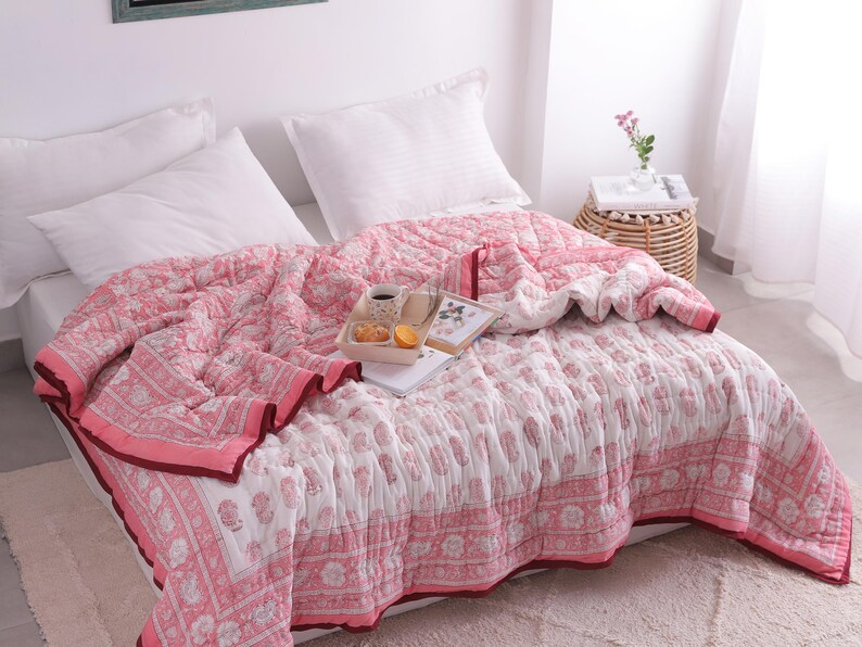 May include: A pink and white floral patterned quilt covers a bed with white pillows. The quilt has a decorative border and is paired with a tray holding a cup, pastries, and an orange slice. A small side table with a vase of flowers is visible.