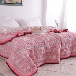 May include: A pink and white floral patterned quilt on a bed with white pillows. The quilt has a dark pink border and a ruffled edge. The bed is on a white rug.
