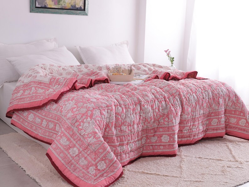 May include: A pink and white floral patterned quilt on a bed with white pillows. The quilt has a dark pink border and a ruffled edge. The bed is on a white rug.