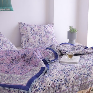 May include: A bed is made with a floral patterned quilt and pillow set in shades of purple and white. The quilt has a dark blue border. A cup of tea and a book are on the bed. A small gray vase with a plant is on the bed.