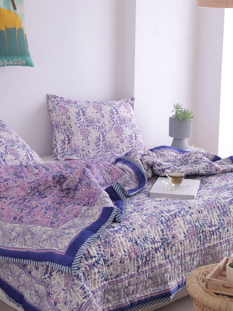May include: A bed is made with a floral patterned quilt and pillow set in shades of purple and white. The quilt has a dark blue border. A cup of tea and a book are on the bed. A small gray vase with a plant is on the bed.