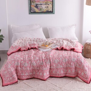 May include: A pink and white floral patterned quilt on a bed with white pillows. The quilt has a ruffled edge and a dark red border. A small woven side table with a vase of flowers is next to the bed.
