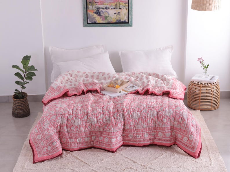 May include: A pink and white floral patterned quilt on a bed with white pillows. The quilt has a ruffled edge and a dark red border. A small woven side table with a vase of flowers is next to the bed.