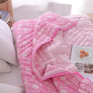 May include: A pink and white floral patterned quilt with a matching pink border is displayed on a bed. The quilt is folded over, revealing a striped pattern on the underside. White pillows and a book with pastries are also visible.