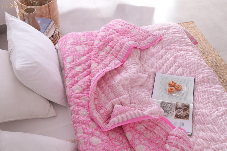 May include: A pink and white floral patterned quilt with a matching pink border is displayed on a bed. The quilt is folded over, revealing a striped pattern on the underside. White pillows and a book with pastries are also visible.