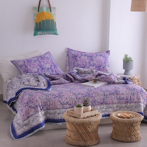 May include: A bed is made with a purple floral patterned quilt and matching pillowcases. The quilt has a white border with a blue trim. Two small woven stools sit in front of the bed. A tote bag with a Nashville skyline hangs on the wall.