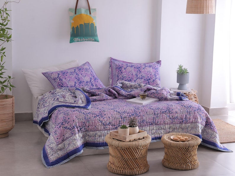 May include: A bed is made with a purple floral patterned quilt and matching pillowcases. The quilt has a white border with a blue trim. Two small woven stools sit in front of the bed. A tote bag with a Nashville skyline hangs on the wall.