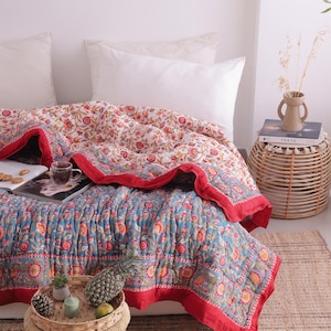 May include: A floral patterned quilt in shades of blue, red, and cream covers a bed. The quilt features a red border and a detailed floral design. A small woven table holds a tray with a pineapple and other decorative items. The bed is made up with white pillows.