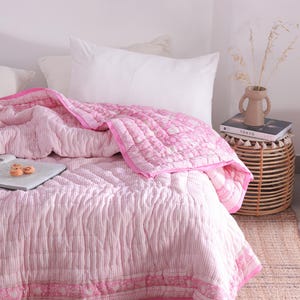 May include: A pink and white striped quilt with a floral border on a bed with white pillows. A small side table holds a vase with dried plants and a book. The room has a neutral colour palette.