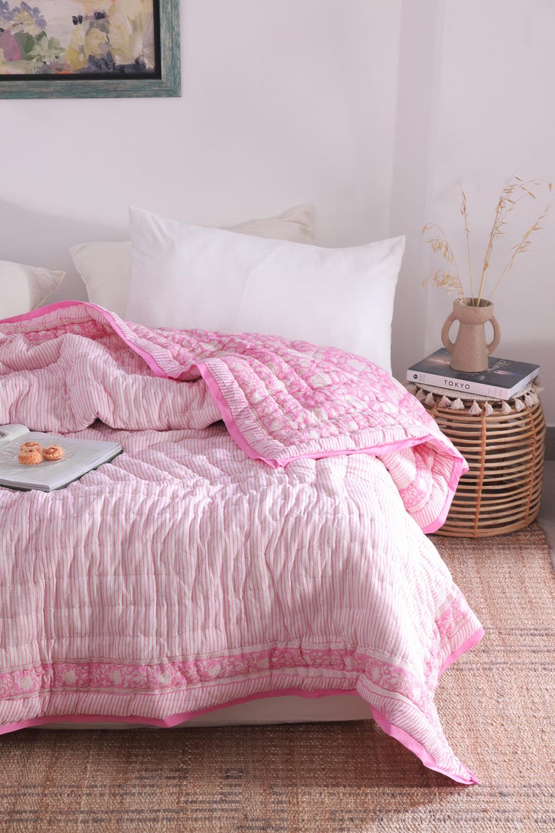May include: A pink and white striped quilt with a floral border on a bed with white pillows. A small side table holds a vase with dried plants and a book. The room has a neutral colour palette.