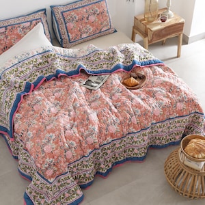 May include: A floral-patterned quilt and matching pillowcases in shades of peach, pink, and blue adorn a bed. The quilt features a detailed floral design with a blue border and pink trim. A wooden side table with a vase and candle is next to the bed.