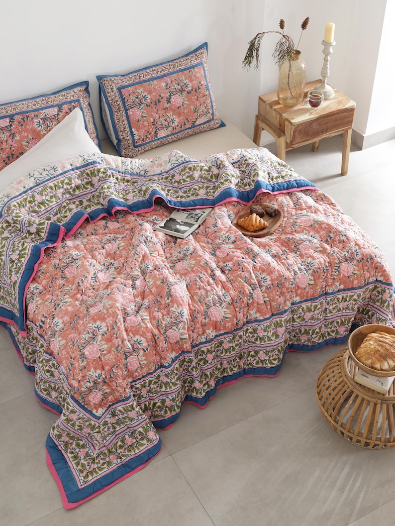 May include: A floral-patterned quilt and matching pillowcases in shades of peach, pink, and blue adorn a bed. The quilt features a detailed floral design with a blue border and pink trim. A wooden side table with a vase and candle is next to the bed.