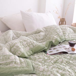 May include: A bed is covered with a light green and white patterned quilt and white pillows. A glass of dark liquid, an open book, and walnuts are on the quilt. The quilt has a green border.
