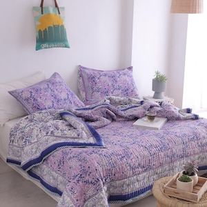 May include: A bed is covered with a floral patterned quilt and matching pillowcases in shades of purple, white, and blue. A tote bag with a Nashville skyline design hangs on the wall. A woven side table holds a tray with small potted plants.