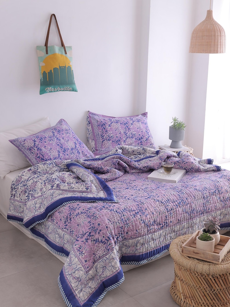 May include: A bed is covered with a floral patterned quilt and matching pillowcases in shades of purple, white, and blue. A tote bag with a Nashville skyline design hangs on the wall. A woven side table holds a tray with small potted plants.