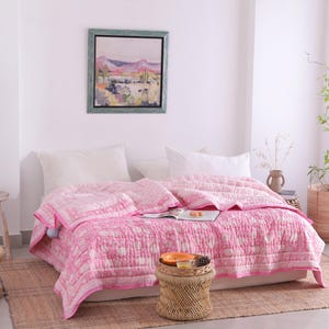 May include: A pink and white patterned quilt on a bed with white pillows. The quilt has a border and is draped over the bed. A small woven stool holds a tray with items. A painting hangs on the wall.