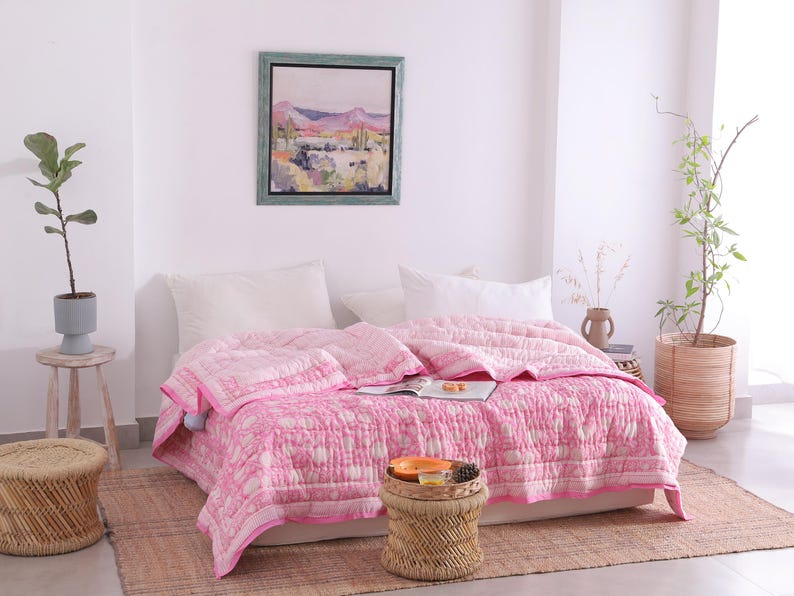 May include: A pink and white patterned quilt on a bed with white pillows. The quilt has a border and is draped over the bed. A small woven stool holds a tray with items. A painting hangs on the wall.
