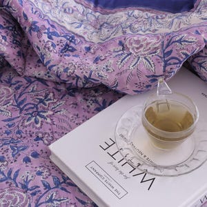 May include: A floral patterned quilt in shades of lavender and blue, with a cup of tea on a saucer resting on a book titled "WHITE". The quilt features a repeating botanical design. The book is from "The White Company".