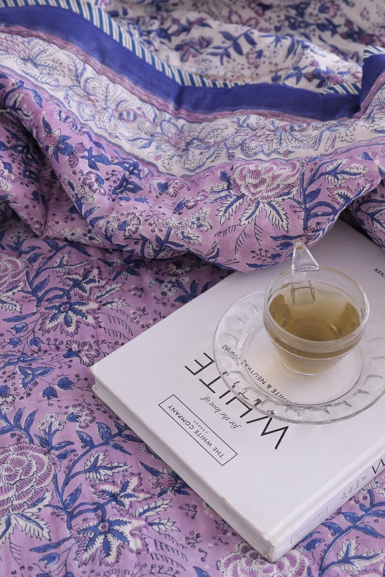 May include: A floral patterned quilt in shades of lavender and blue, with a cup of tea on a saucer resting on a book titled "WHITE". The quilt features a repeating botanical design. The book is from "The White Company".