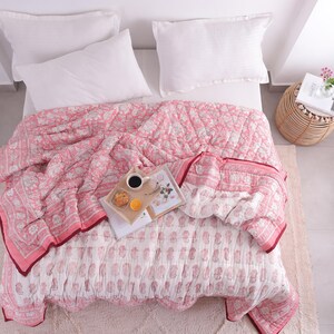 May include: A bed is covered with a pink and white floral patterned quilt. Two white pillows rest on the bed. A tray with coffee, a pastry, and an orange slice sits on the quilt. A small side table holds a book and flowers.