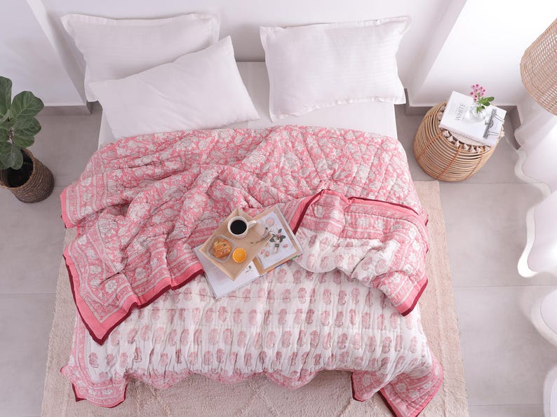May include: A bed is covered with a pink and white floral patterned quilt. Two white pillows rest on the bed. A tray with coffee, a pastry, and an orange slice sits on the quilt. A small side table holds a book and flowers.