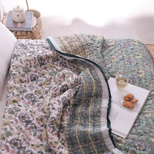 Op de afbeelding: Een bloemmotief quilt in groen, roze en wit, met een contrasterende groene rand. De quilt ligt op een bed met witte kussens. Een boek, een glas thee en koekjes liggen op de quilt.