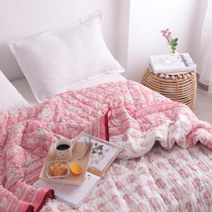 May include: A bed is made with white pillows and a pink floral patterned quilt. A wooden tray holds a cup of coffee, a pastry, and an orange slice. A small woven side table holds a vase of flowers and a book.